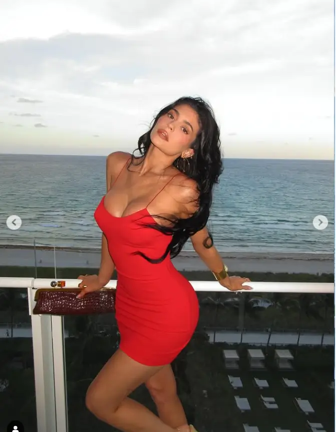 Her choice of attire, a daring red dress, captivates attention with its snug fit. This design accentuates her curvaceous figure, while the deep neckline showcases considerable cleavage.